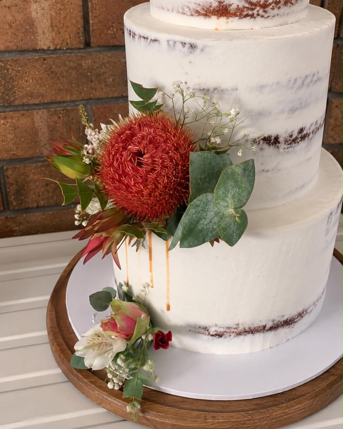 Close up of three tier semi-naked wedding cake with caramel drip, large red pincushion protea, eucalyptus and pink and white flowers on a wooden cake board